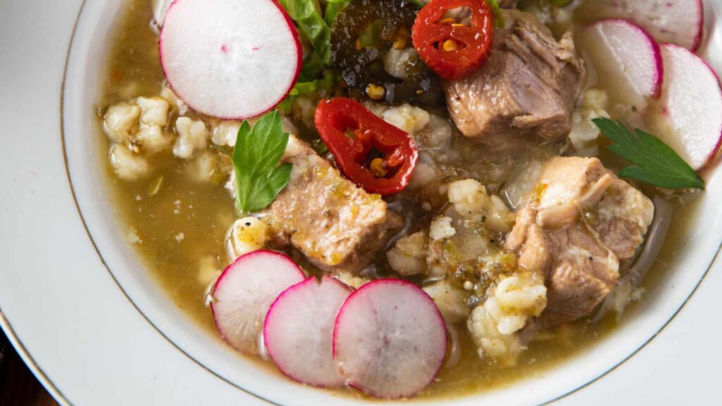 A closeup of pork pozole verde in a white bowl with slices of radishes placed for garnish.