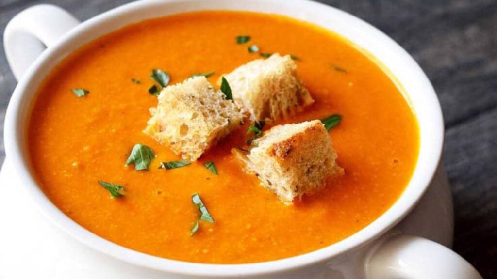 A white crock filled with Instant Pot tomato soup, garnished with three bread crumbs and fresh herbs.