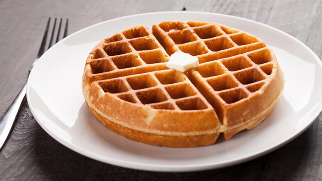 A single round waffle on a white plate, garnished with a pat of butter. A fork lays to the left of the plate.
