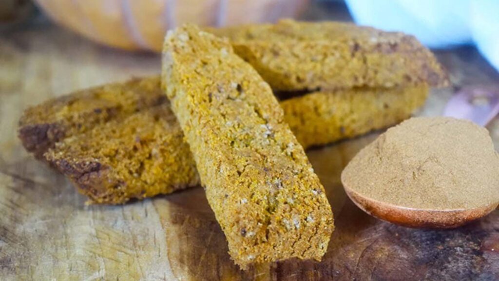 Gluten-Free Pumpkin Biscotti laying on a wooden surface next to a heaping tablespoon of pumpkin pie spice.