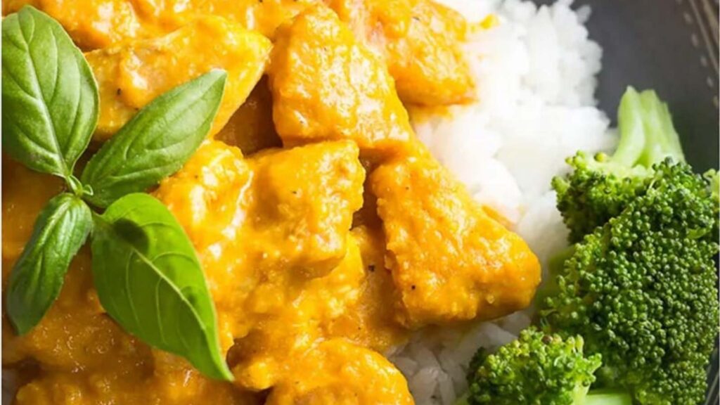 A closeup of Pumpkin Chicken Curry on a bed of white rice with a side of broccoli and a garnish of fresh basil leaves.