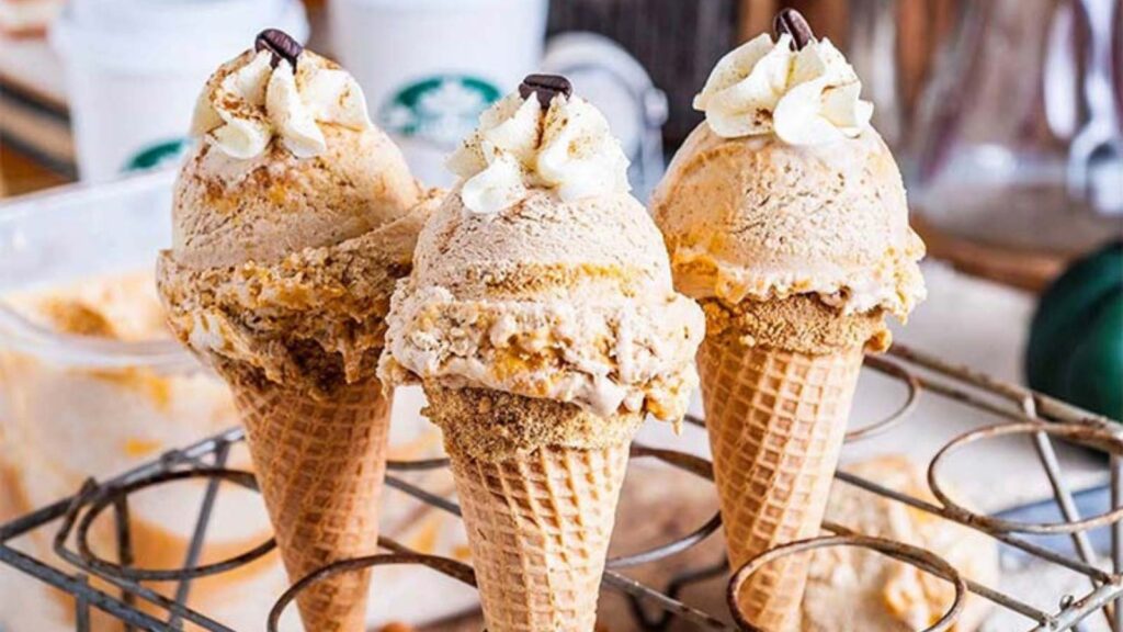 Three ice cream cones in a cone holder with a single scoop of No Churn Pumpkin Spice Latte Ice Cream on each, and each one is topped with a dollop of whipped cream and a single coffee bean.