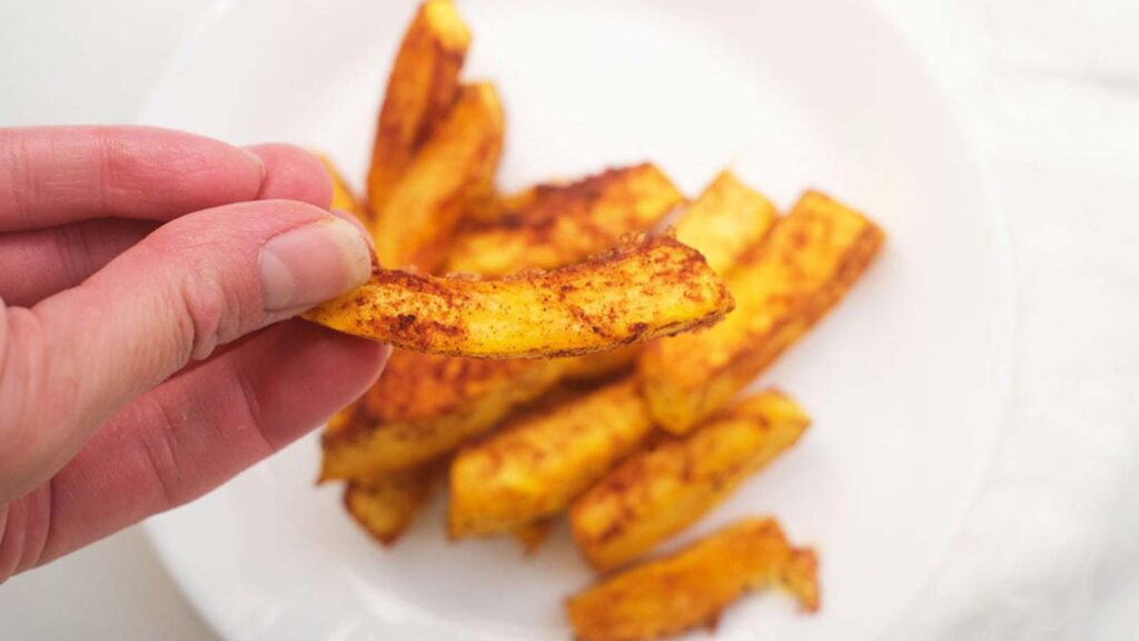 A hand holds up a single Pumpkin Fry over a plate filled with them.