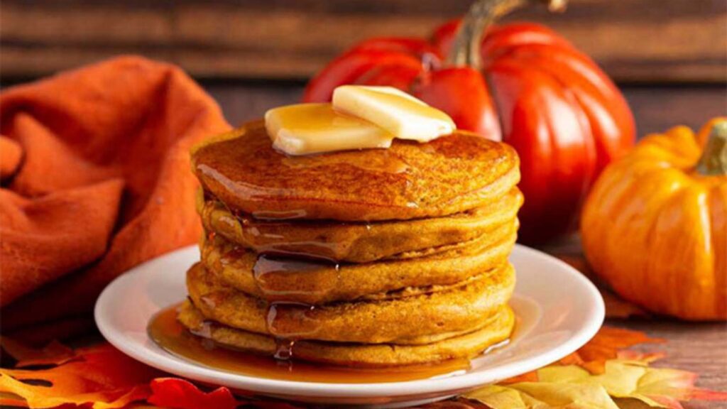A stack of Pumpkin Pancakes on a white plate with two pats of butter on top and maple syrup dripping down the front.