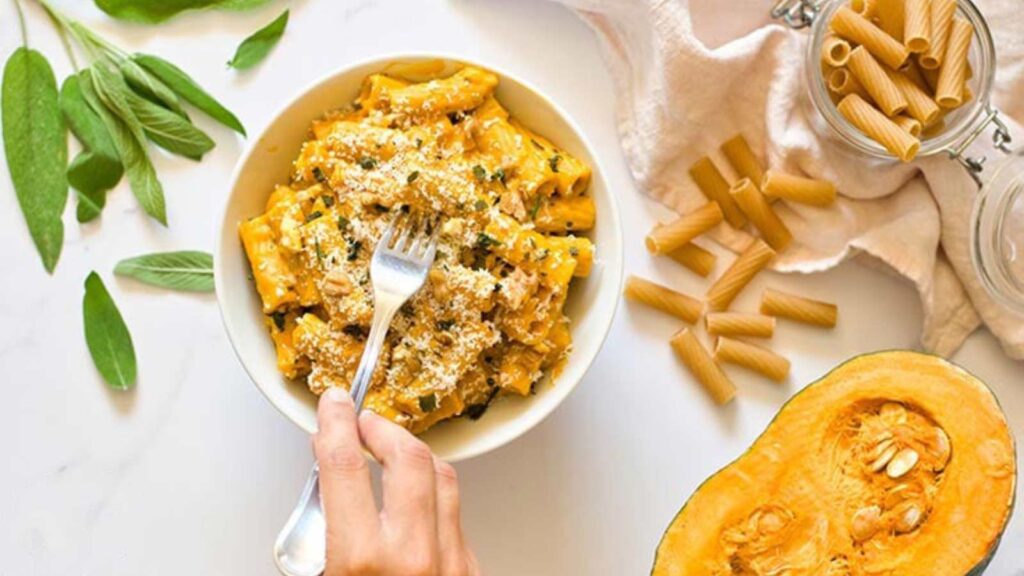A female hand holding a fork picks up some Pumpkin Pasta out of a white bowl.