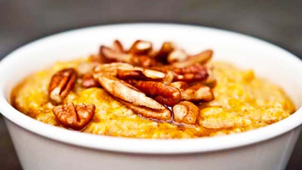 A white bowl holds a serving of pumpkin pie oatmeal and is topped with pecans and maple syrup.