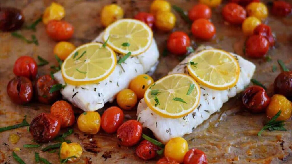 Two roasted cod filets on a roasting pan with small tomatoes all around them.