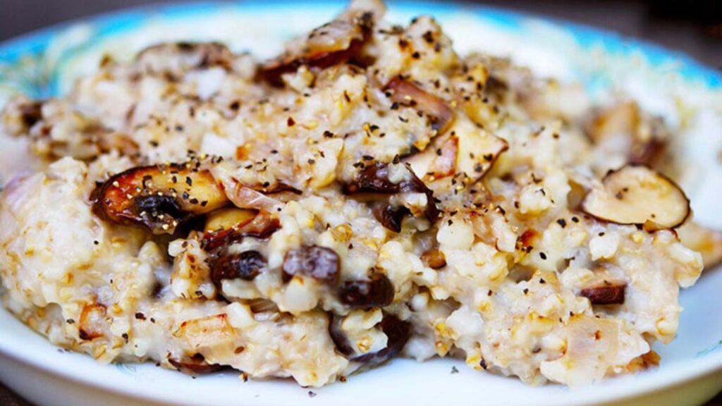 A bowl of Savory Oatmeal sits on a dark background. You can see the golden color of the saut&eacute;ed mushrooms mixed into the oats.