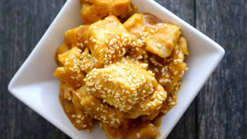 An overhead view of a square, ceramic, white dish holding a serving of sesame chicken garnished with sesame seeds.