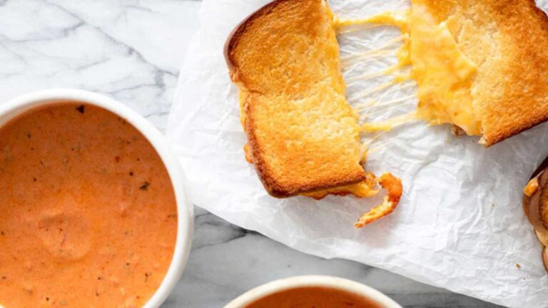 A Sheet Pan Grilled Cheese Sandwich cut in half and laying on parchment next to two bowls of tomato soup.