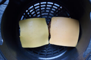 Two turkey patties in an air fryer basket with slices of cheese place on them.