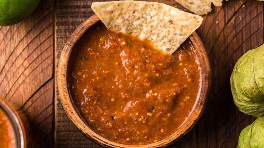 A wood bowl filled with salsa and one corn chip resting in the dip.