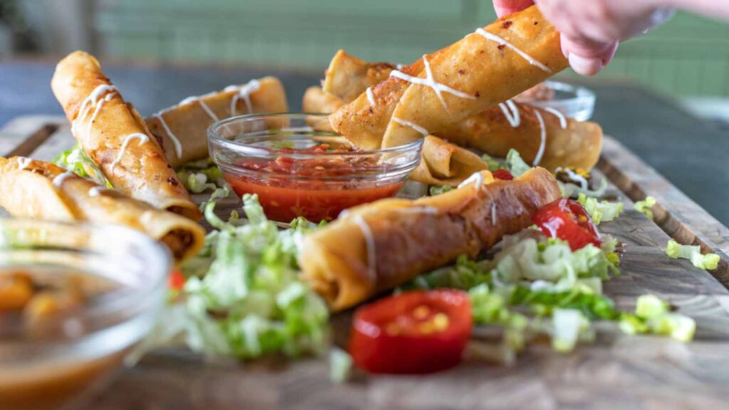 A cutting board with venison taquitos. A hand dips one into a bowl of red sauce.