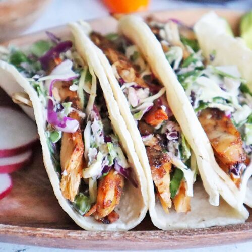 Jerk chicken tacos with slaw and radishes on a wooden plate.