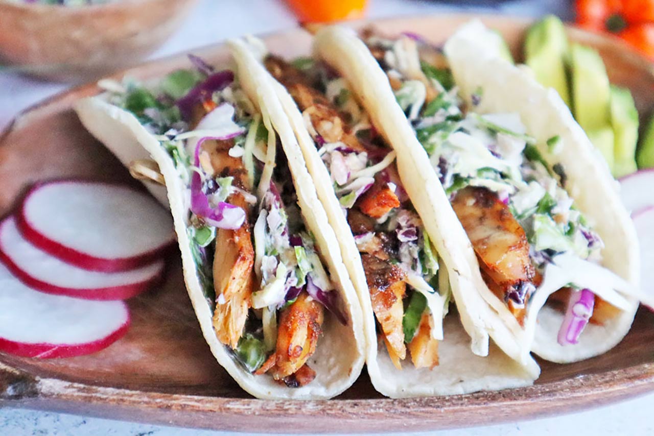 Jerk chicken tacos with slaw and radishes on a wooden plate.