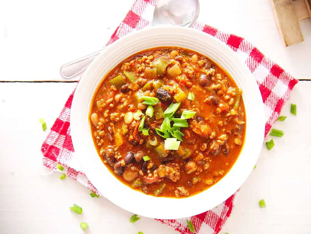 An overhead view of a white bowl filled with beef chili, sitting on a placemat on a table with a spoon.