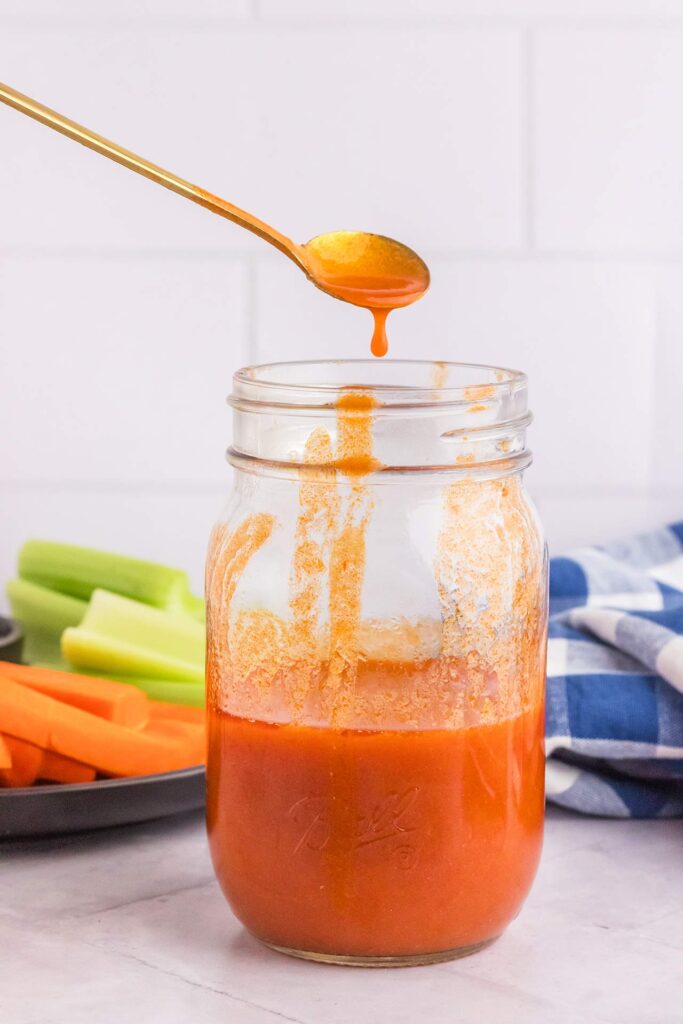 A spoon drips some buffalo sauce into a jar full of it.