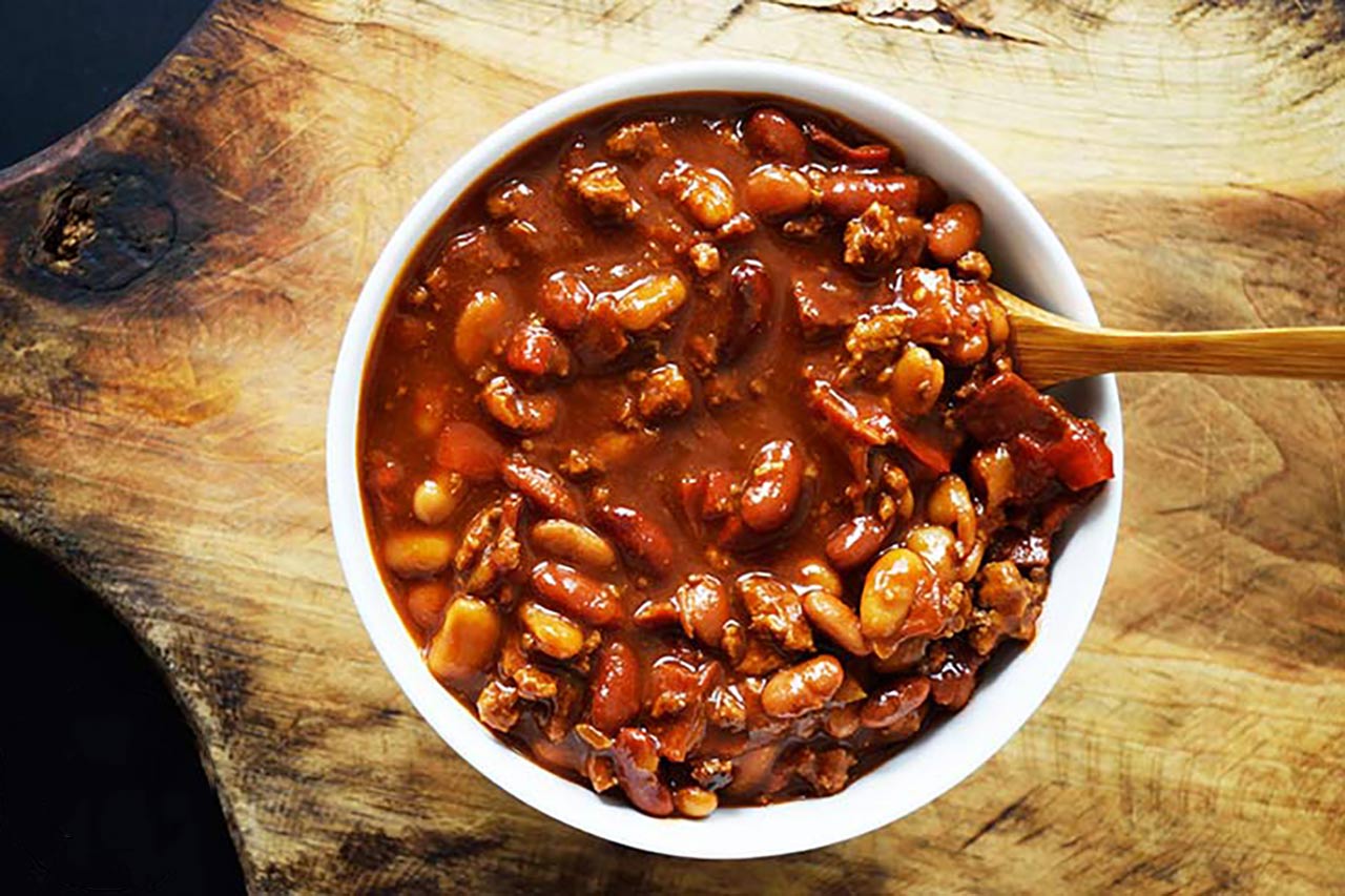 An overhead view looking down into a white bowl filled with cowboy beans. A wooden spoon rest in the beans.