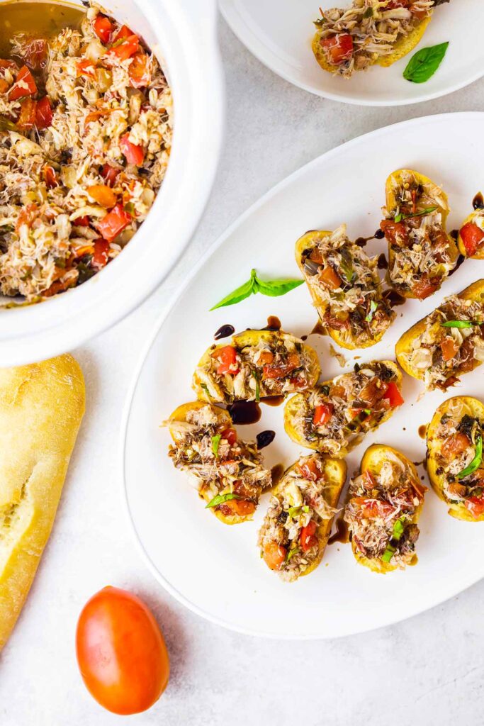 An overhead view of a platter of crab crostini, a crock filled with crab mix, a Roma tomato and a baguette.
