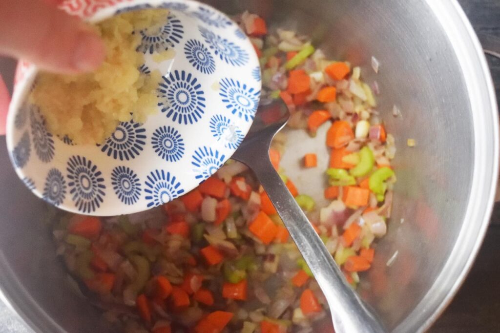 Adding pressed garlic to a soup pot of chopped vegetables.