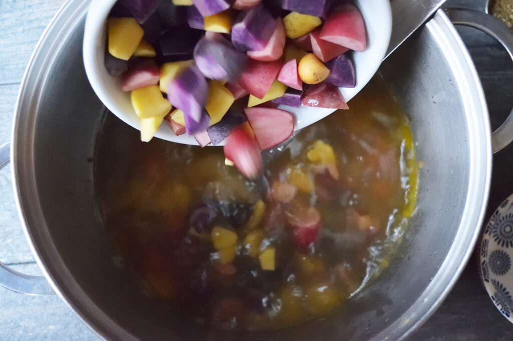 Adding cut potato chunks to a soup pot of stock and chopped veggies.