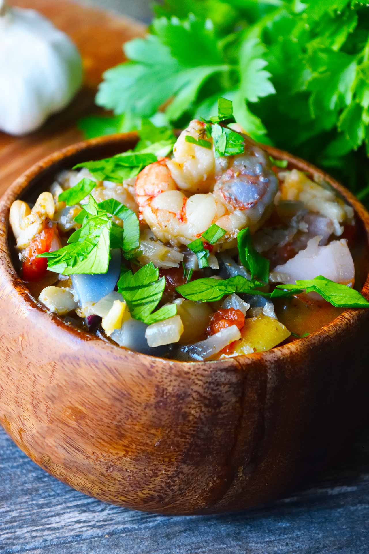 A wood bowl filled with Chunky Fish Stew. A bunch of fresh parsley sits behind it with a head of garlic.