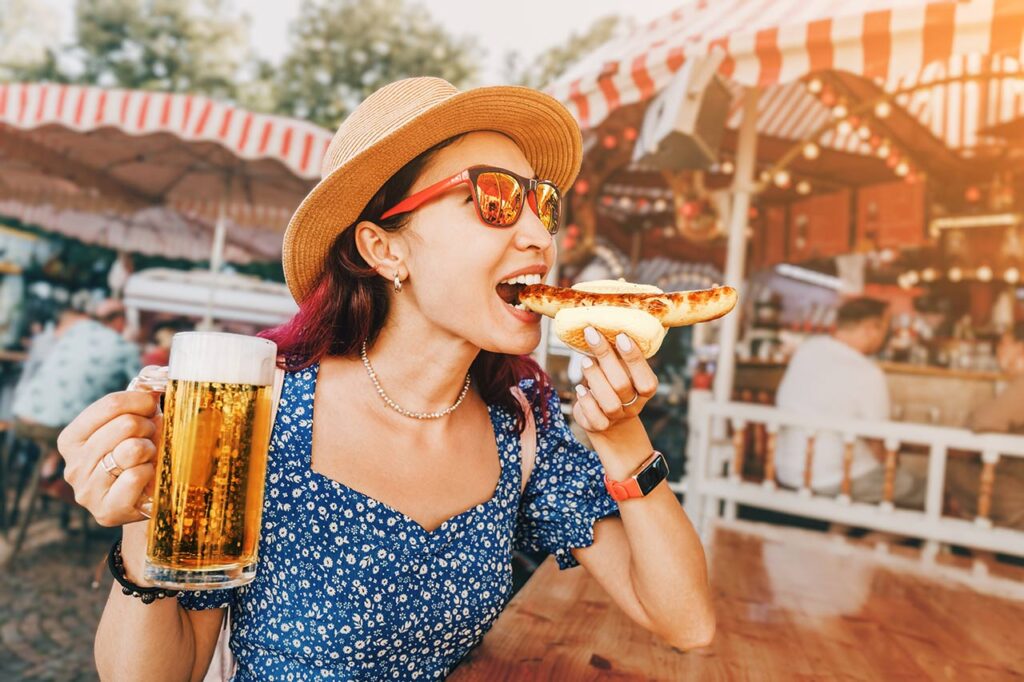 Happy girl drinking beer and eating traditional german bratwurst - hotdog at fun fair and street food festival.