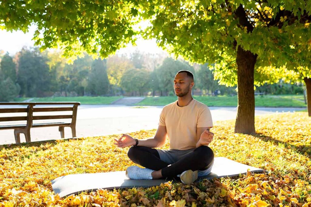 A man sits on a yoga mat, meditating in a park.