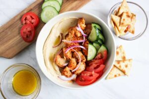 An overhead view of a Mediterranean Bowl With Chicken, cucumbers and tomatoes. Pita chips sit to the side.