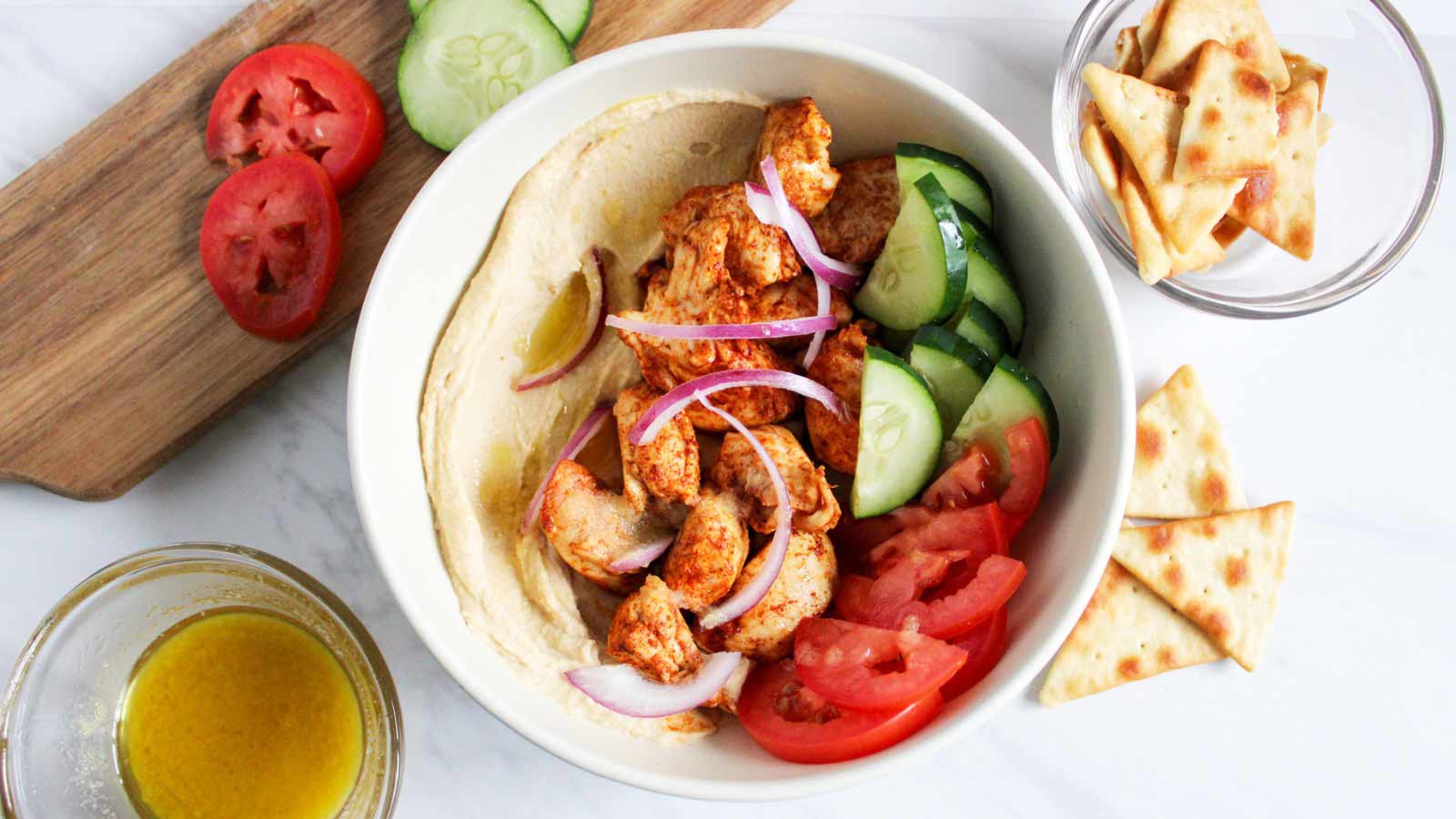 An overhead view of a Mediterranean Bowl With Chicken, cucumbers and tomatoes. Pita chips sit to the side.