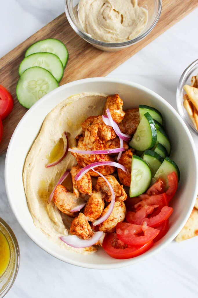 An overhead view of a Mediterranean Bowl With Chicken. Cucumbers and tomatoes rest to the side of the bowl on a cutting board.