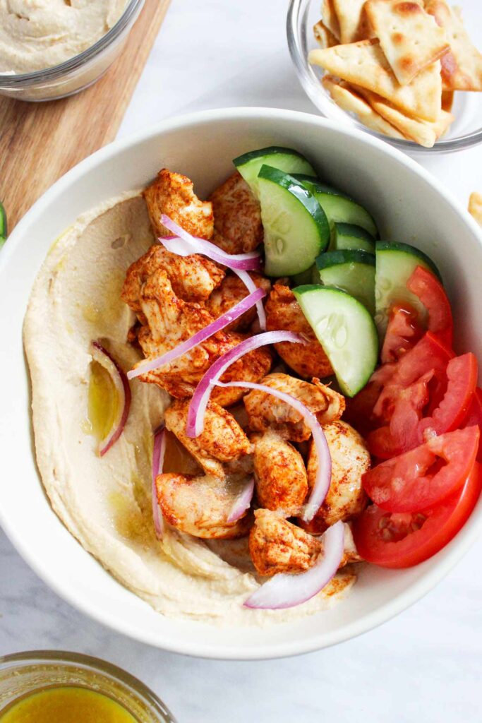 A white bowl from overhead filled with this Mediterranean Bowl Recipe. Hummus, chicken, cucumbers, tomatoes and onions fill the bowl.