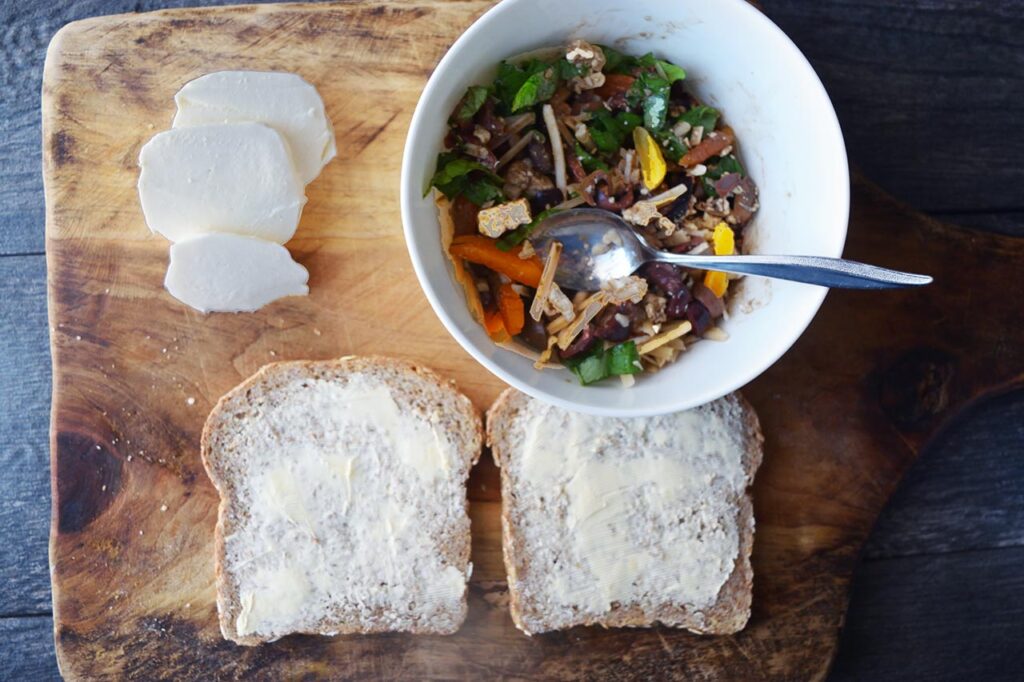 Sandwich filling, sliced mozzarella, and buttered slices of whole grain bread on a cutting board.