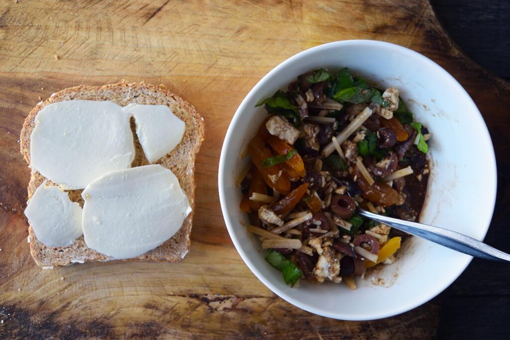 Sliced mozzarella pieces on a piece of whole grain bread next to a bowl of sandwich filling.