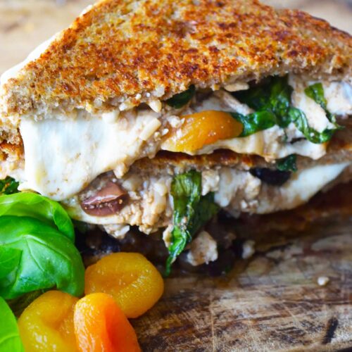 A stacked Mediterranean Sandwich on a cutting board with fresh basil leaves and dried apricots.