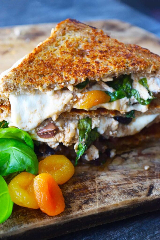 A stacked Mediterranean Sandwich on a cutting board with fresh basil leaves and dried apricots.