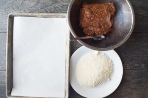 A setup work space for making Date And Oat Energy Balls. A parchment-lined baking sheet, a bowl of dough and a plate of shredded coconut.
