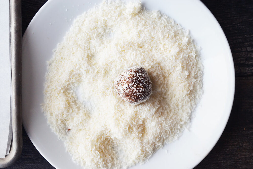 A Date And Oat Energy Ball rolled in coconut and laying on a plate full of coconut shreds.