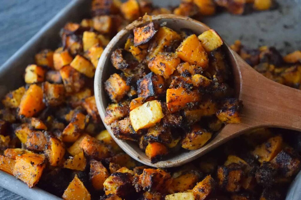 A wooden scoop of Roasted Butternut Squash lays on a sheet pan full of more squash.