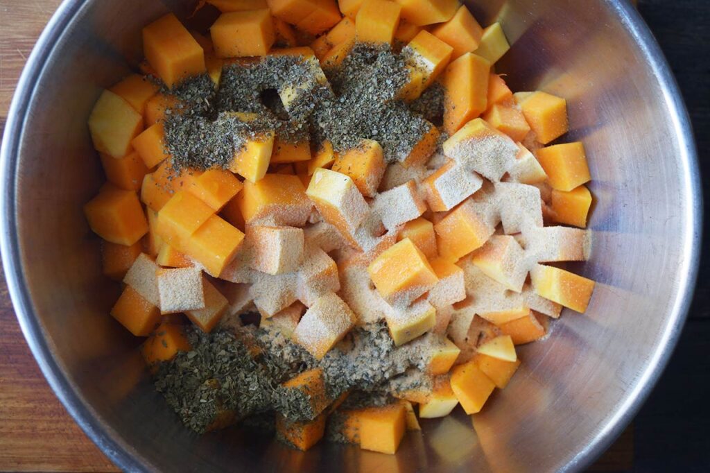 Herbs and spices added to butternut squash cubes in a mixing bowl.