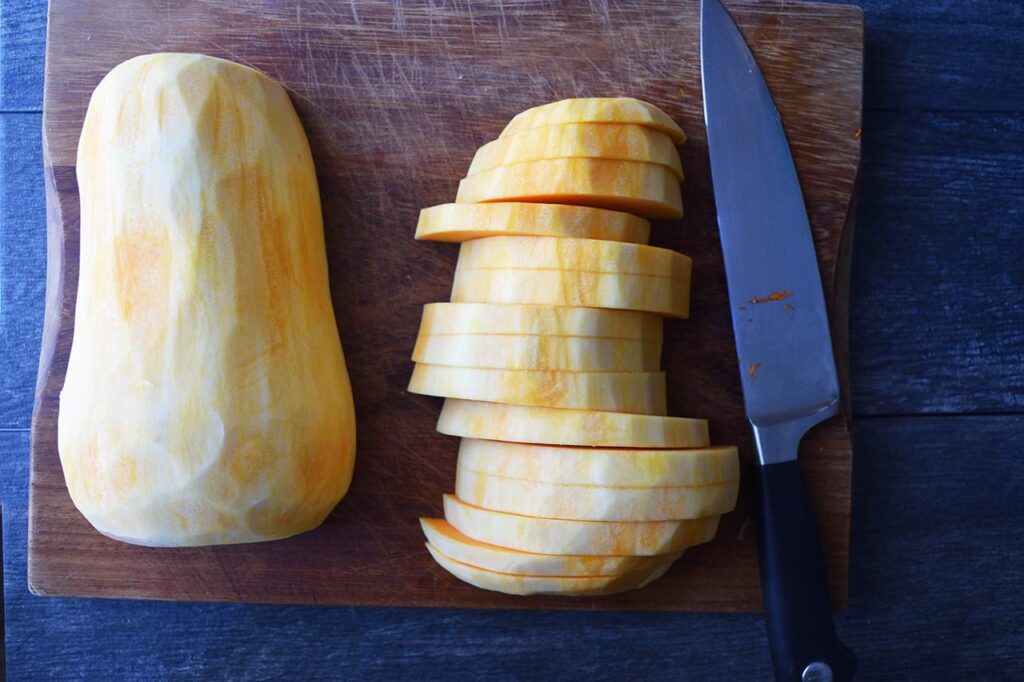 Two halves of a butternut squash on a cutting board. One half is sliced into half-inch slices.