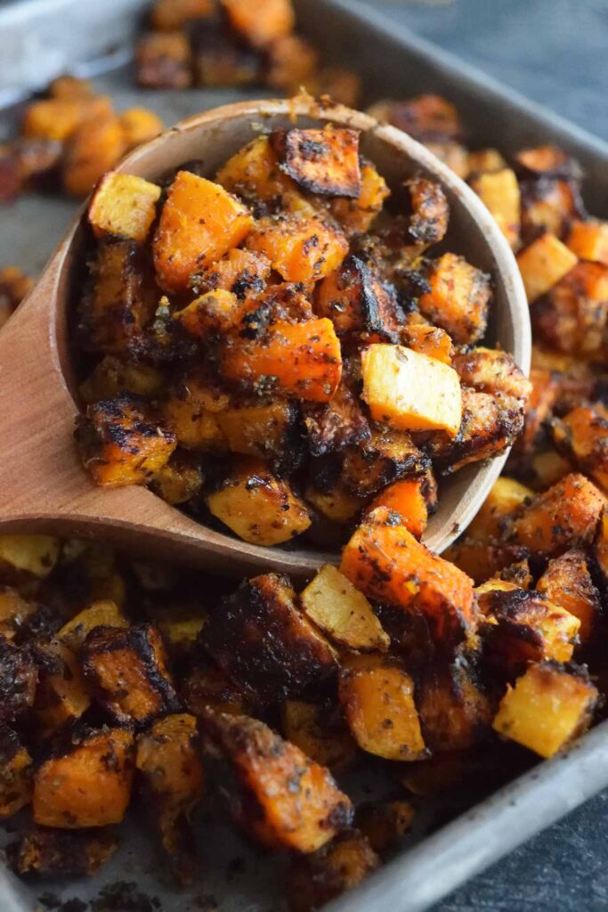 A side view of a wooden scoop filled with Roasted Butternut Squash, laying on a sheet pan full of squash.