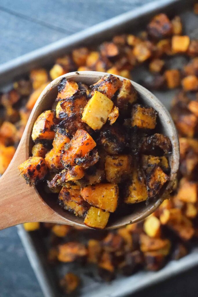 A wooden scoop lifts a scoopful of Roasted Butternut Squash off of a sheet pan.