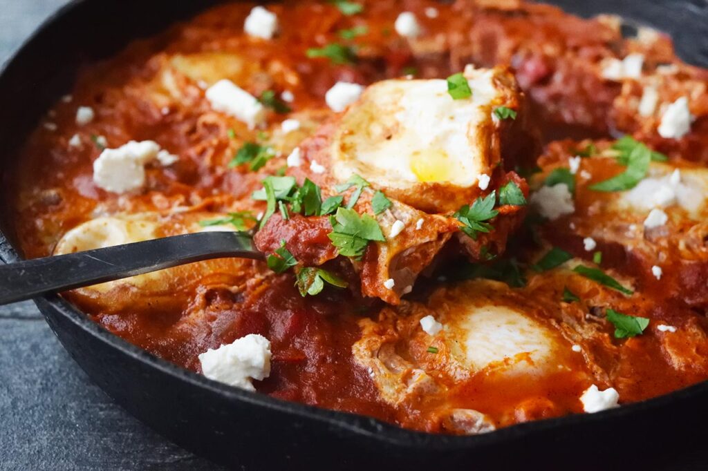 A serving spoon rests in a cast iron skillet, with a spoonful of Shakshuka.