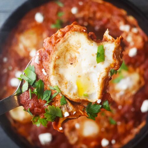 A serving spoon lifts a spoonful of Shakshuka above the cast iron pan it was cooked in.