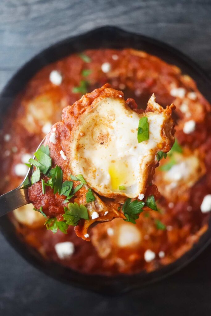 A serving spoon lifts a spoonful of Shakshuka above the cast iron pan it was cooked in.