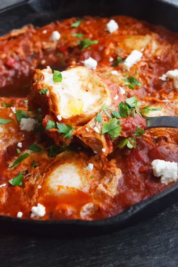 Side view of a serving spoon lifting a spoonful of Shakshuka out of a cast iron skillet.