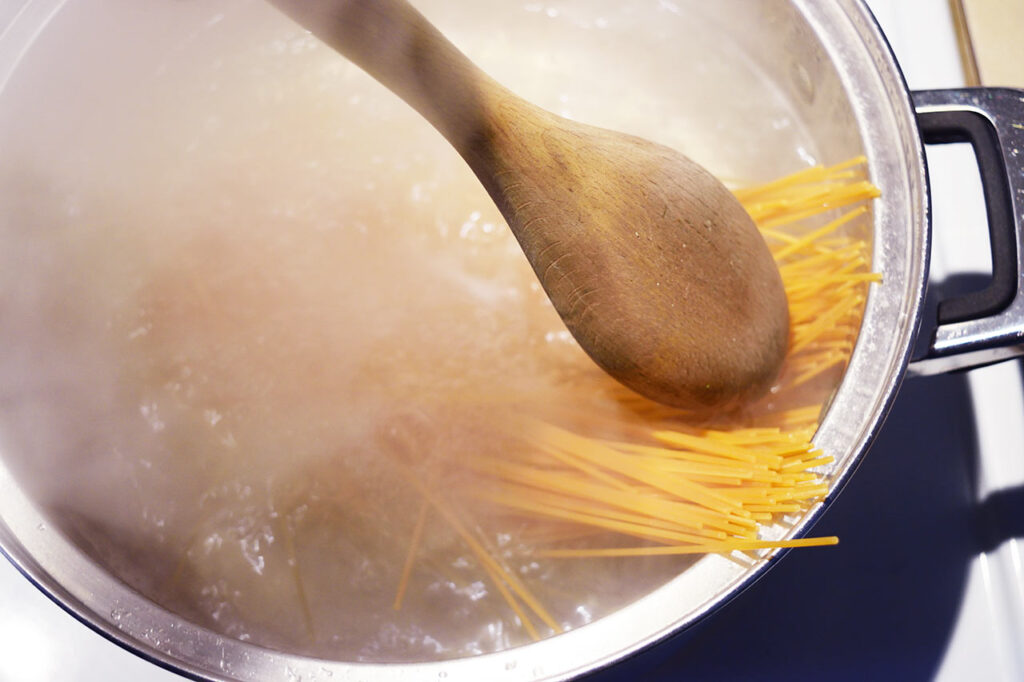 A wooden spoon pushing raw pasta down into salted pasta water.