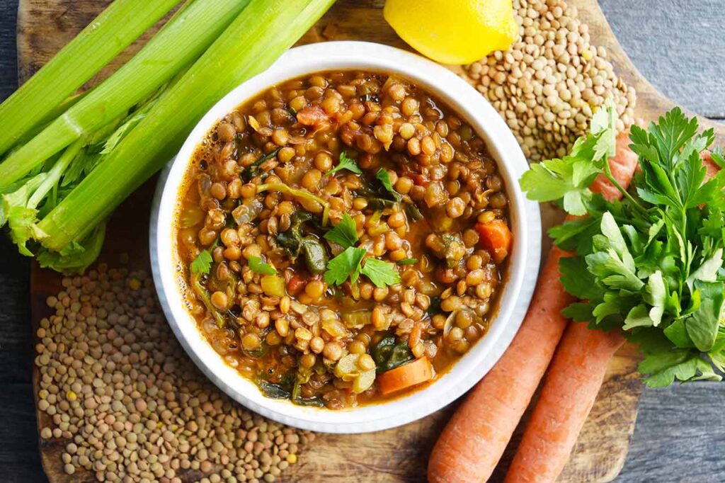 An overhead view of a white bowl filled with Spinach Lentil Soup, surrounded by produce and dried lentils.