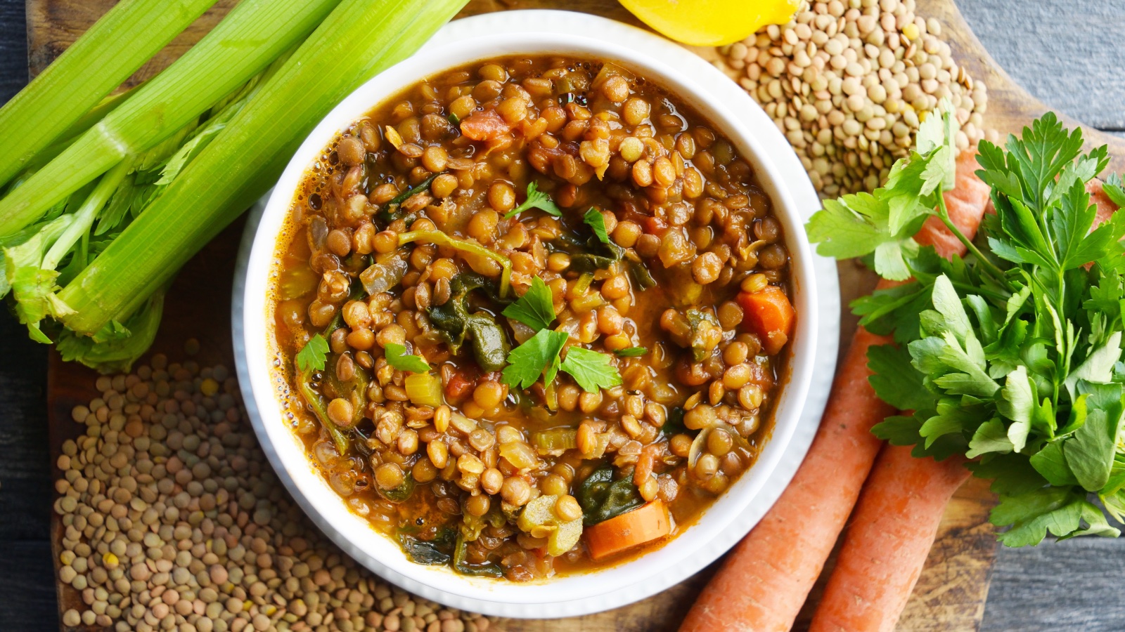 An overhead view of a white bowl filled with Spinach Lentil Soup, surrounded by produce and dried lentils.